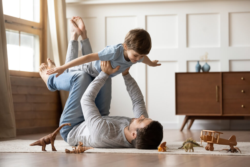 Happy young father lying on floor in living room hold fly with little preschooler son engaged in funny game together, loving dad relax playing with small boy child, enjoy family