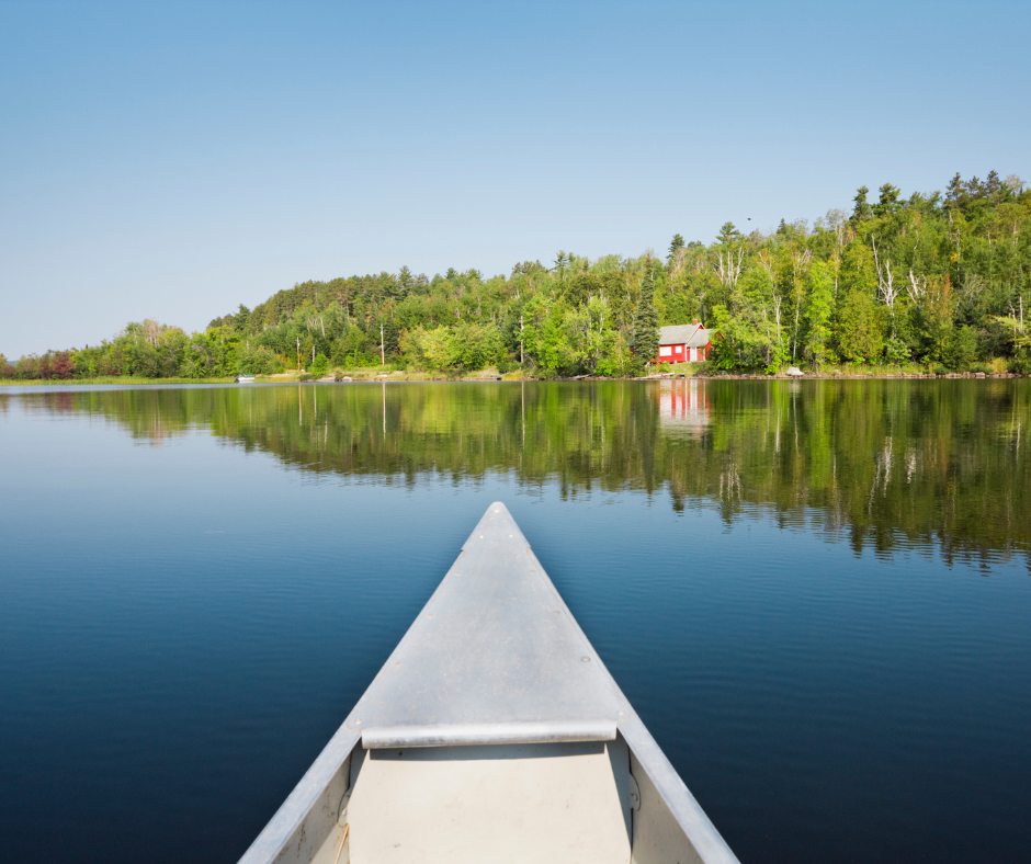 Canoe heading toward a forested shoreline with a red cabin on a calm lake under a clear blue sky.