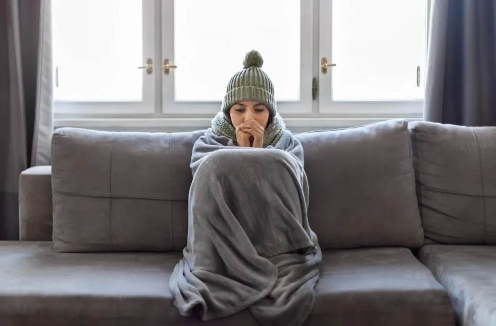 Cold Home. Young Woman Covered With Blanket Freezing On Couch In Living Room, Millennial Female Wearing Knitted Scarf And Hat Indoors Warming Hands With Her Breath,