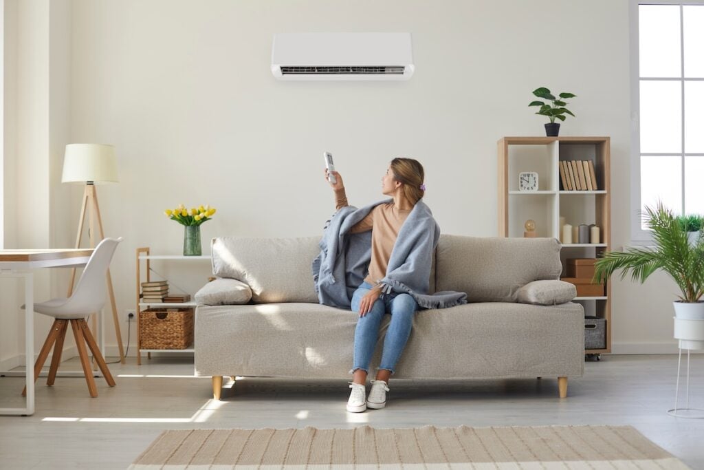 Woman who's sitting on sofa under warm plaid in living room switches off her air conditioner on wall.