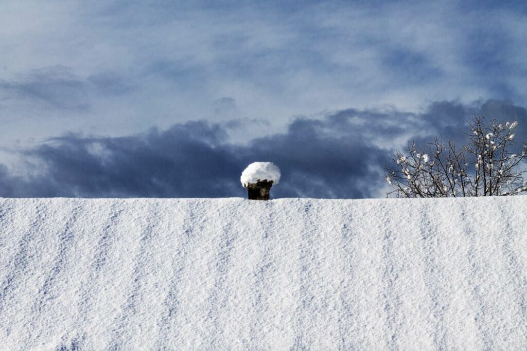 a large roof of a house with a small chimney covered with snow against a dark overcast cloudy sky