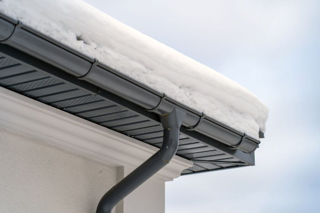 Corner of house with new roof made of gray metal tiles and gutter covered with thick layer of snow in winter