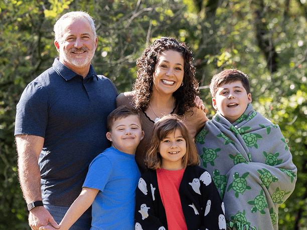 Family of five posing outdoors, smiling at the camera in a sunny park setting.