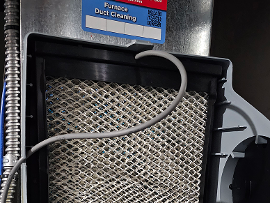 Close-up of an HVAC furnace intake with a metal mesh filter, a gray hose draped over the top, and a blue label above reading 'Furnace Duct Cleaning'