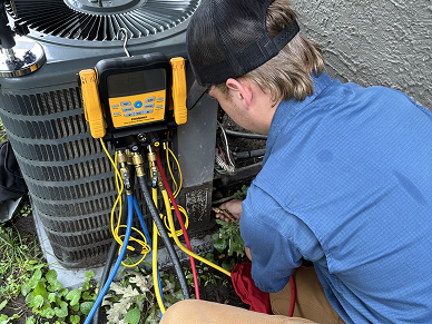 HVAC technician crouches beside an outdoor air conditioner, using a yellow manifold gauge to diagnose the system.