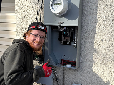 Electrician with a headlamp and red glove working on an outdoor electrical meter box mounted on a textured wall.