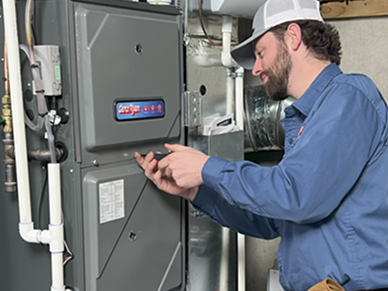 HVAC technician in a blue shirt and cap adjusting a furnace control panel in a mechanical room
