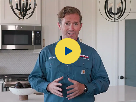 Presenter in a teal work shirt speaks to the camera in a bright modern kitchen; a play button is overlaid on the image indicating a video thumbnail.